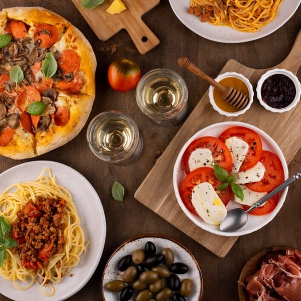 Overhead view of a G's Pizzeria pepperoni and mushroom pizza, spaghetti, caprese salad, olives, and wine.