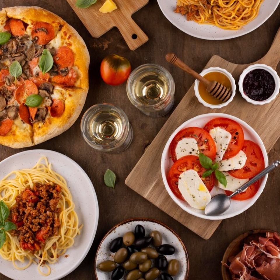 Overhead view of a G's Pizzeria pepperoni and mushroom pizza, spaghetti, caprese salad, olives, and wine.